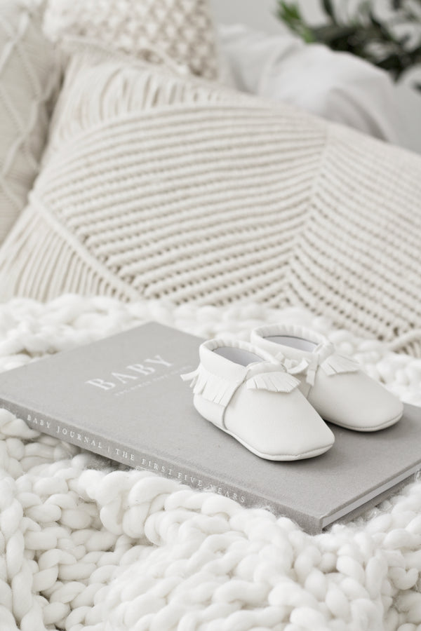 Soft grey linen baby journal titled “BABY – The First Five Years” resting on a chunky white knit blanket with tiny white baby moccasins on top and textured cream cushions in the background.