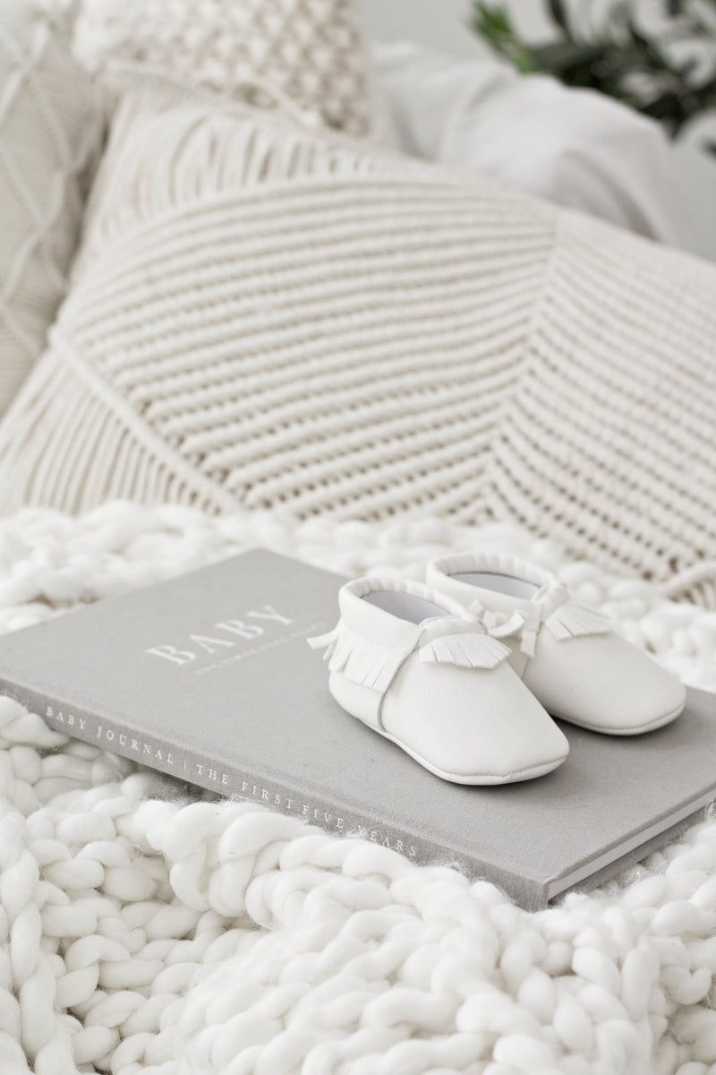 Soft grey linen baby journal titled “BABY – The First Five Years” resting on a chunky white knit blanket with tiny white baby moccasins on top and textured cream cushions in the background.