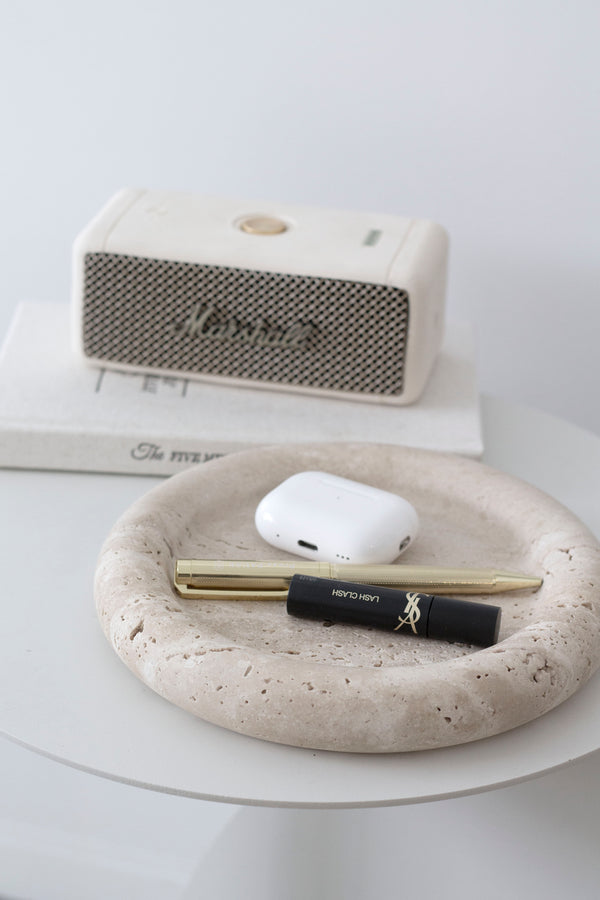 Bay Round Travertine Tray in light stone finish styled with YSL mascara, gold pen, and AirPods pro on a side table.