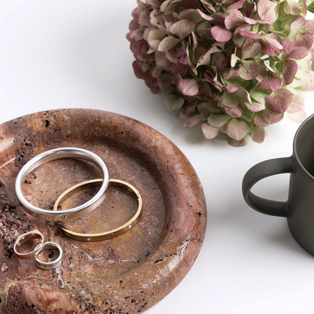 Round tray in a darker red-brown travertine holding bangles and rings beside dried hydrangea and a mug