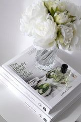 The Beauty Chef Gut Guide styled with white peonies in a glass vase and a perfume bottle on a round white table.