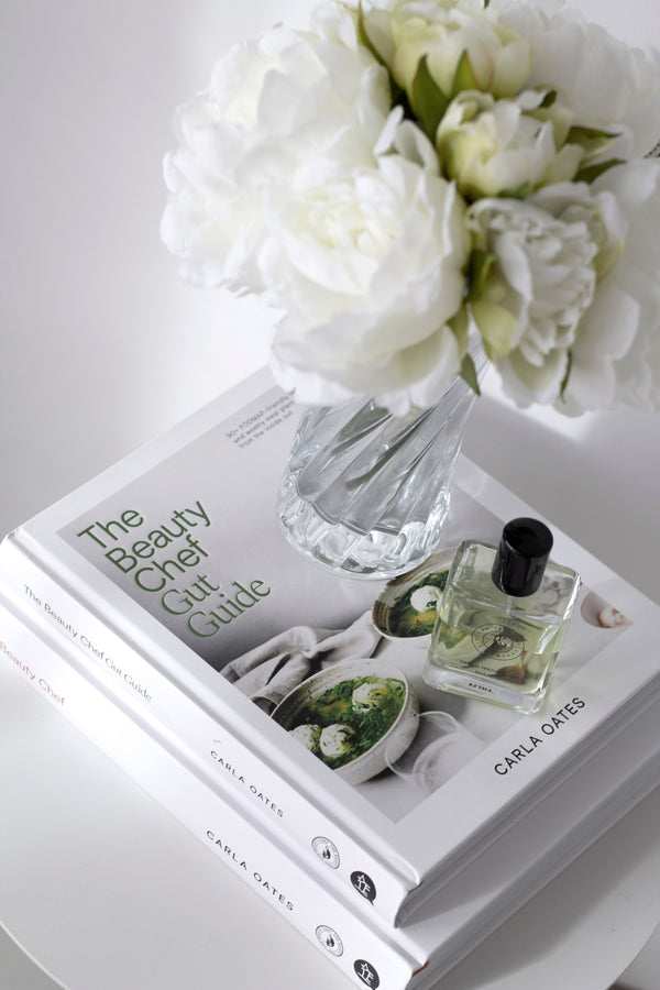 The Beauty Chef Gut Guide styled with white peonies in a glass vase and a perfume bottle on a round white table.