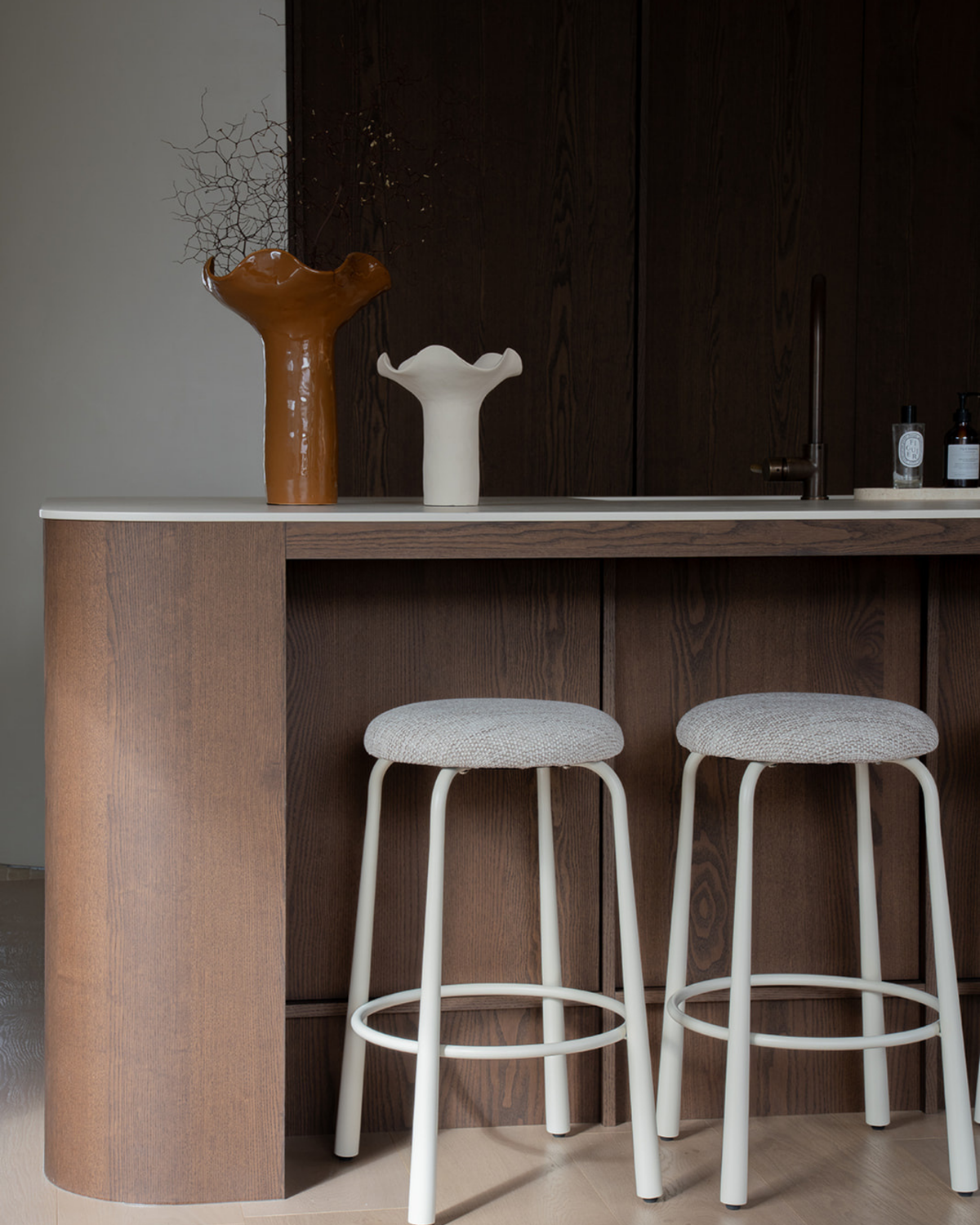 Mud vase with dried branches beside the Ivory vase on a kitchen bench above two upholstered stools