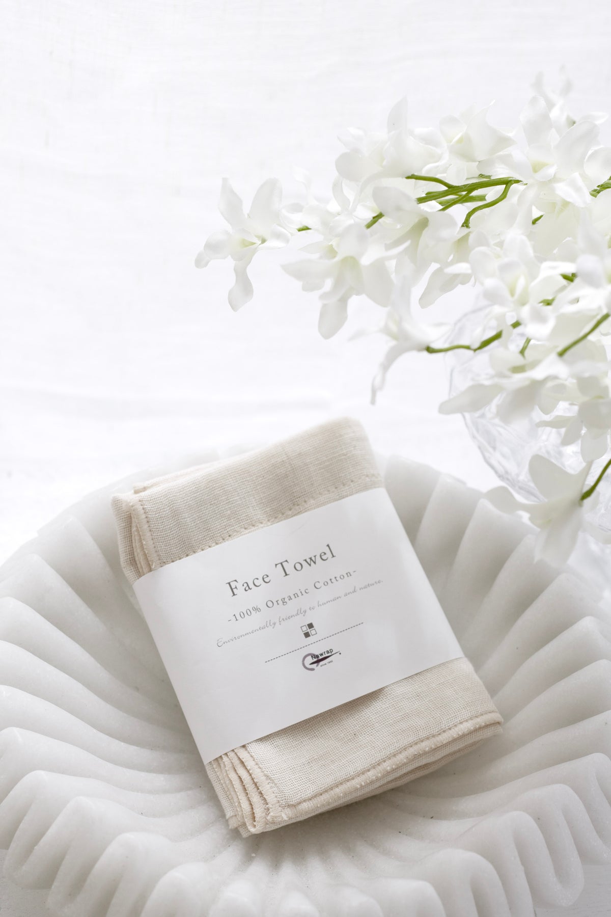 Cream face towel in its paper band in a white bowl beside white blossom
