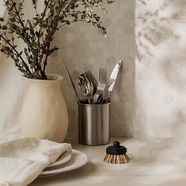 Dish brush and utensils in a stone crock beside a vase of foliage