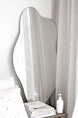 Organic blob-shaped full-length mirror leaning by soft linen curtains in a neutral, Scandi-minimal bedroom.