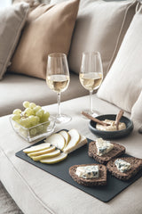 Black rectangular chopping board with cheese, sliced pear, and rye bread, styled with grapes, dip, and two glasses of white wine on a beige sofa