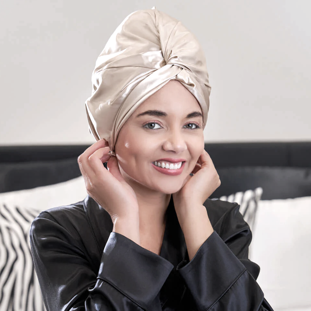 Woman wearing the champagne mulberry silk sleep bonnet, sitting on a bed in a satin robe