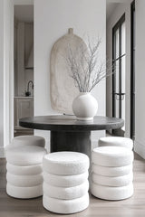 A sculptural white bouclé ottoman with stacked tier design, styled around a dark round table in a minimalist interior with soft natural light.
