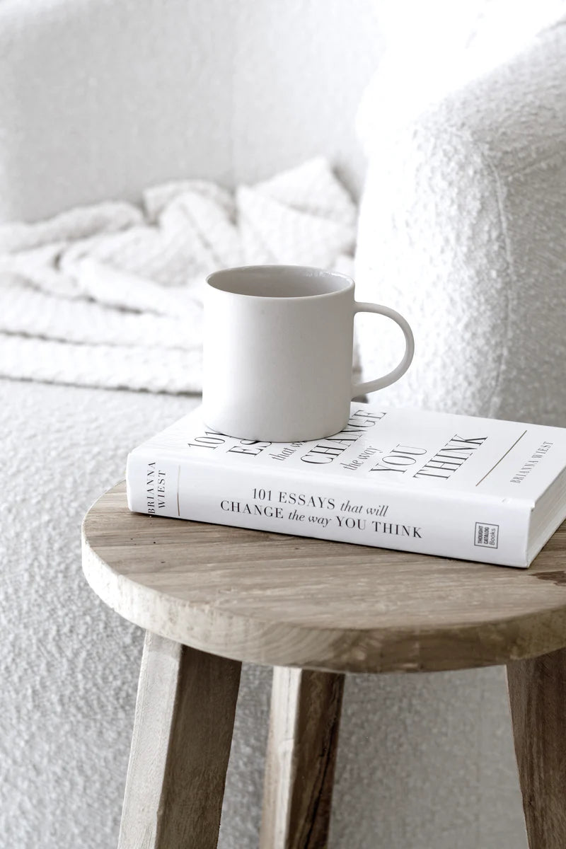 The 101 Essays paperback on a rustic timber stool with a pale stoneware mug resting on top