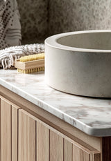 Close-up of a natural nail brush with beechwood handle and Tampico bristles resting beside a concrete basin on a marble vanity.