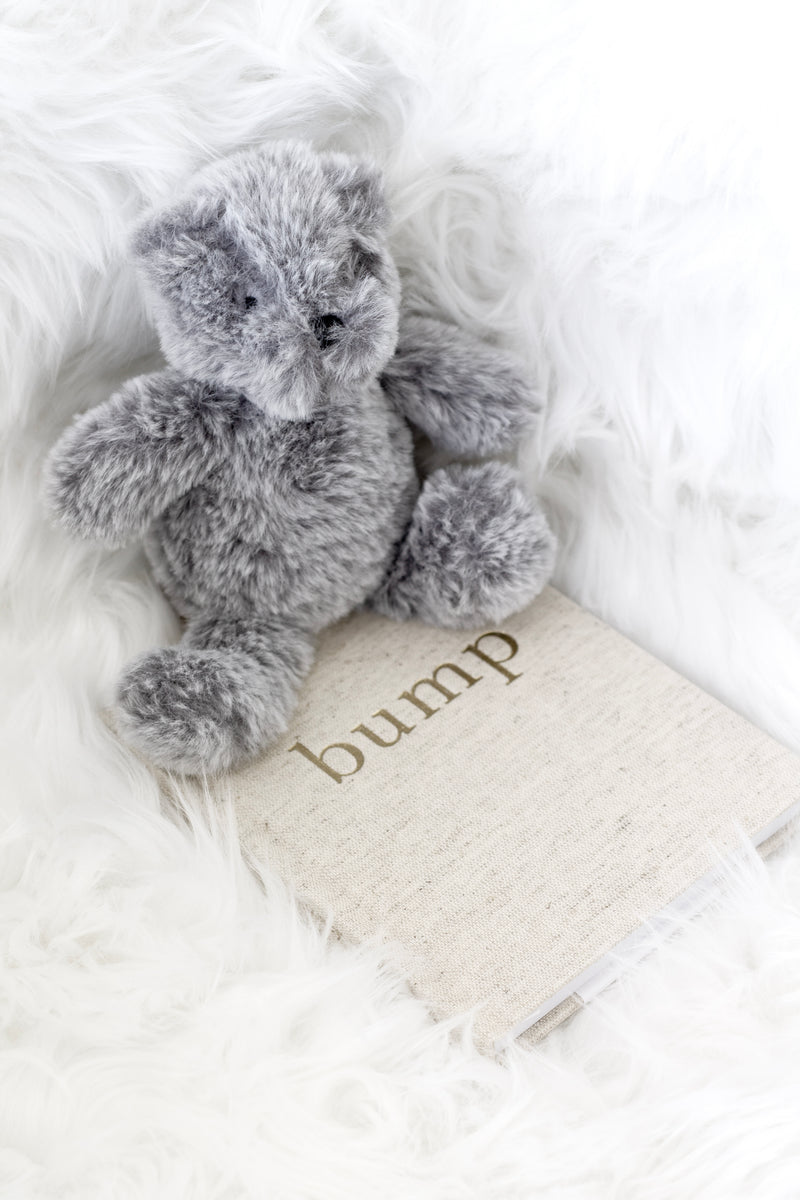 Soft grey plush teddy bear resting on a linen Bump pregnancy journal, styled on a fluffy white faux fur background.