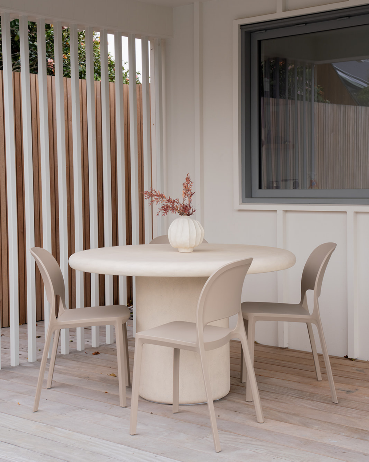 Nude Cali chairs around a round pale table on a covered deck with white battens and a ribbed vase of dried stems