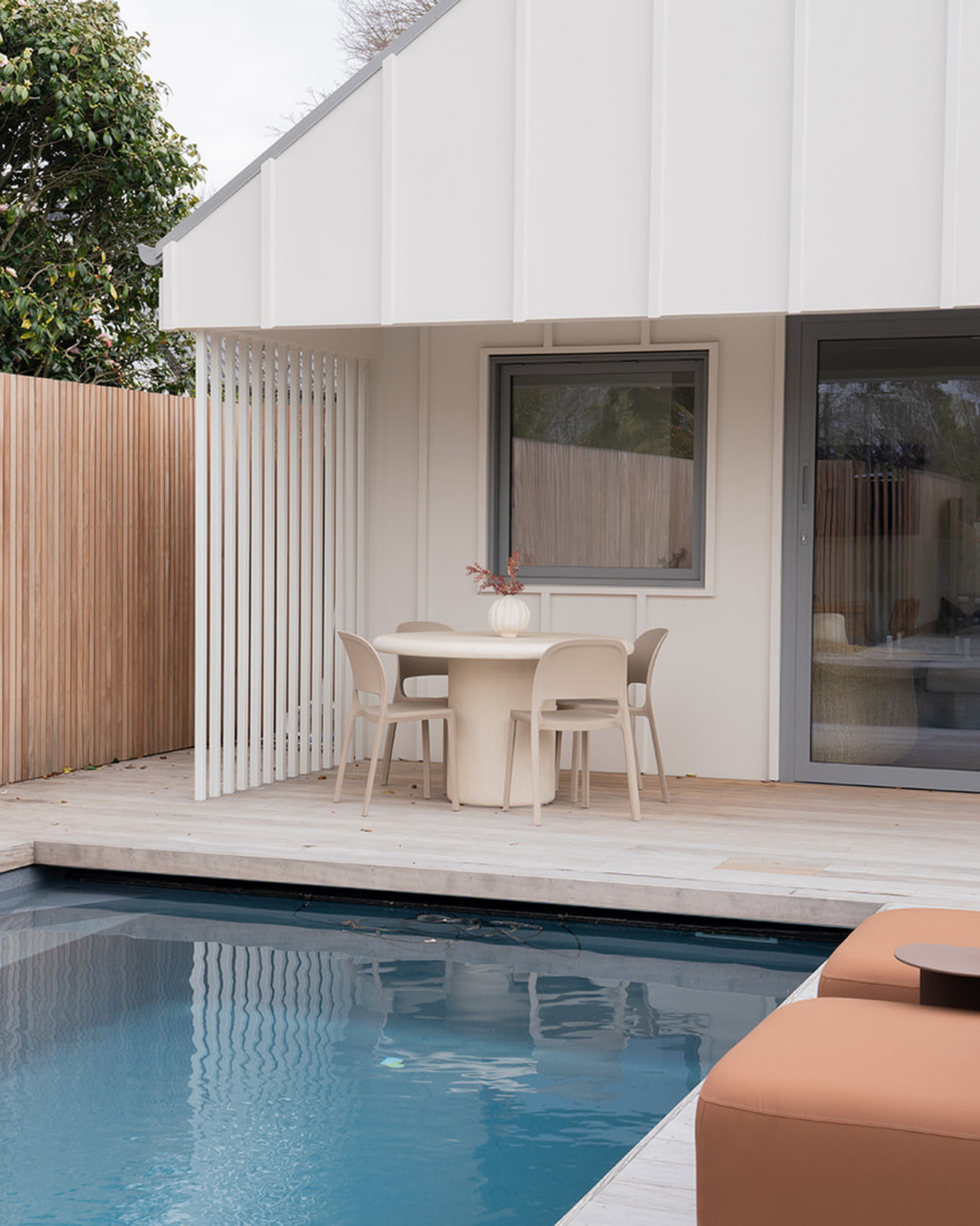 Four Nude Cali chairs around a round table on a pale timber deck beside a pool and white board and batten wall