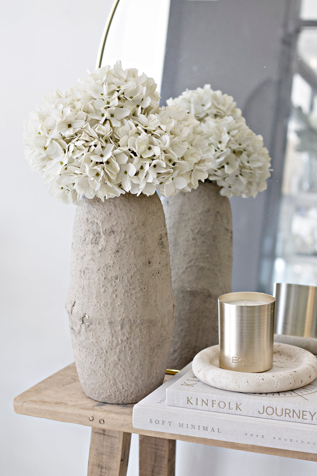 White hydrangeas in two textured vases on a timber stool beside a round mirror and brass candles
