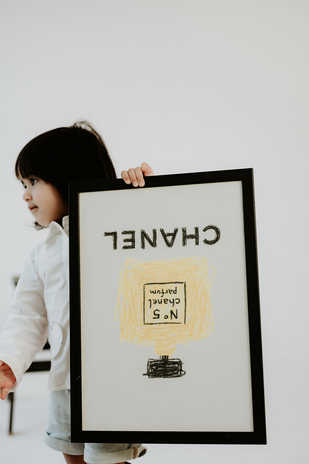 Child holding the Chanel Parfum print in a black frame upside down against a white wall
