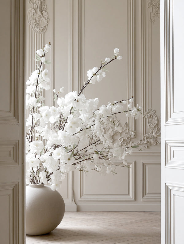Tall white faux cherry blossom stems arranged in a large neutral floor vase in an elegant room with ornate wall panels.