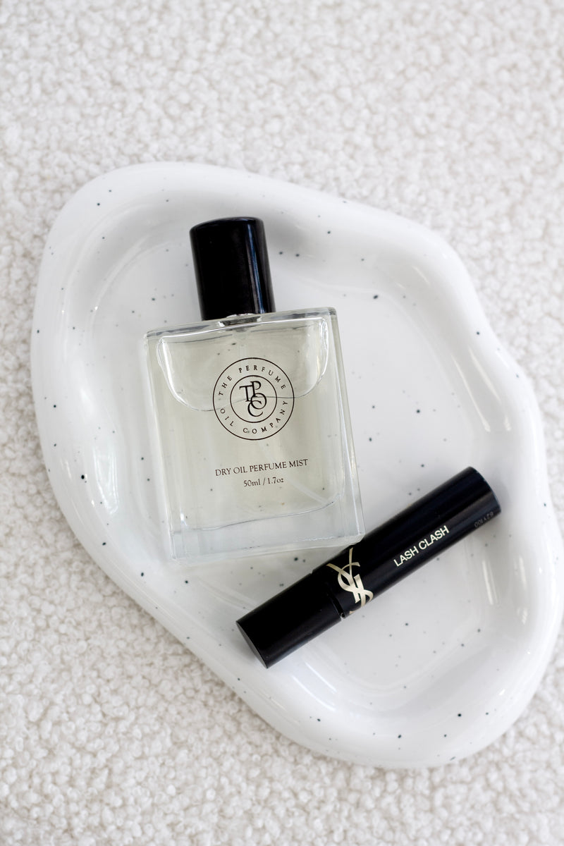 Speckled white cloud-shaped ceramic tray holding a square glass perfume bottle and a black mascara tube, styled on a soft textured cream surface.