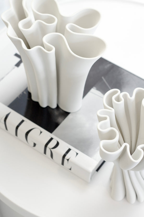 White sculptural 3D-printed vase with flowing wavy edges styled on a monochrome coffee table book in a minimal modern space.