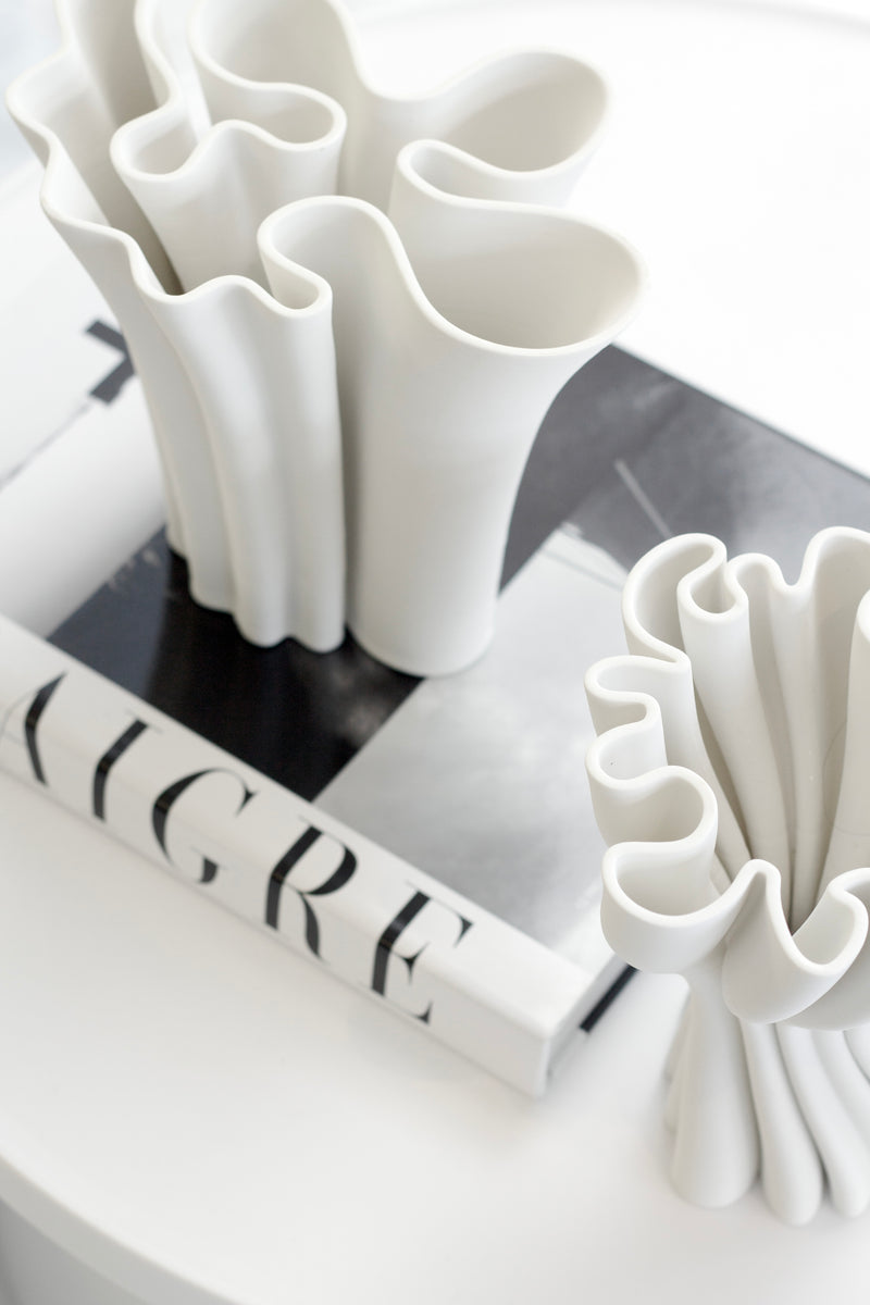 White sculptural 3D-printed vase with flowing wavy edges styled on a monochrome coffee table book in a minimal modern space.