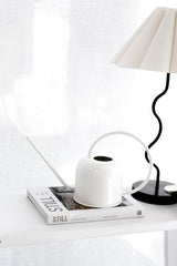 Matte white watering can with sculptural loop handle styled on top of a neutral coffee table book, beside a black curved lamp and soft linen backdrop.