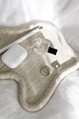 Cream Vanille perfume oil bottle styled on a sculptural travertine tray with rings, AirPods case and a white pen on white bedding in soft sunlight