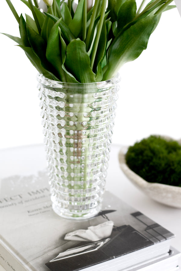 Cut glass vase with white tulips styled on a book beside a moss-filled sculptural bowl on a white table.