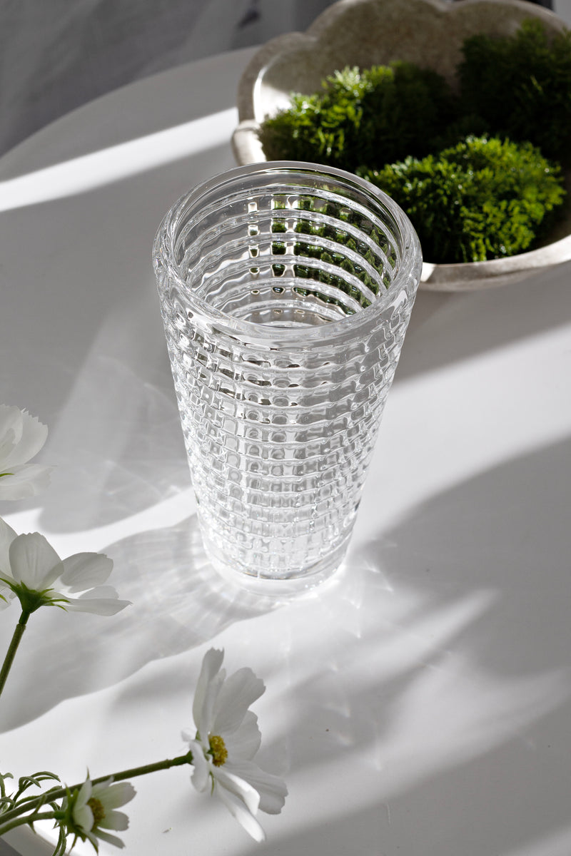 Clear textured cut-glass vase catching the light on a white round table, styled with white flowers in the foreground and a shallow bowl of moss in the background.
