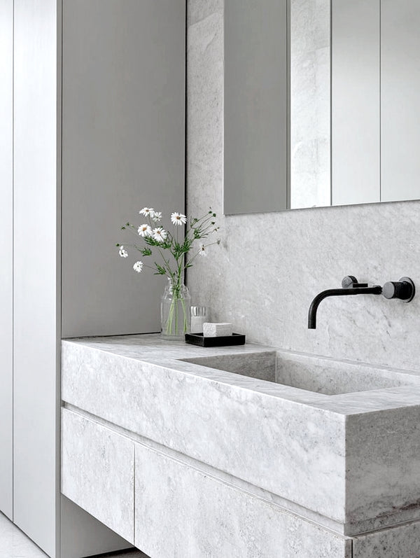 Minimalist bathroom with a large rectangular marble sink, matte black wall-mounted faucet, and a glass vase of white wildflowers styled on the vanity.