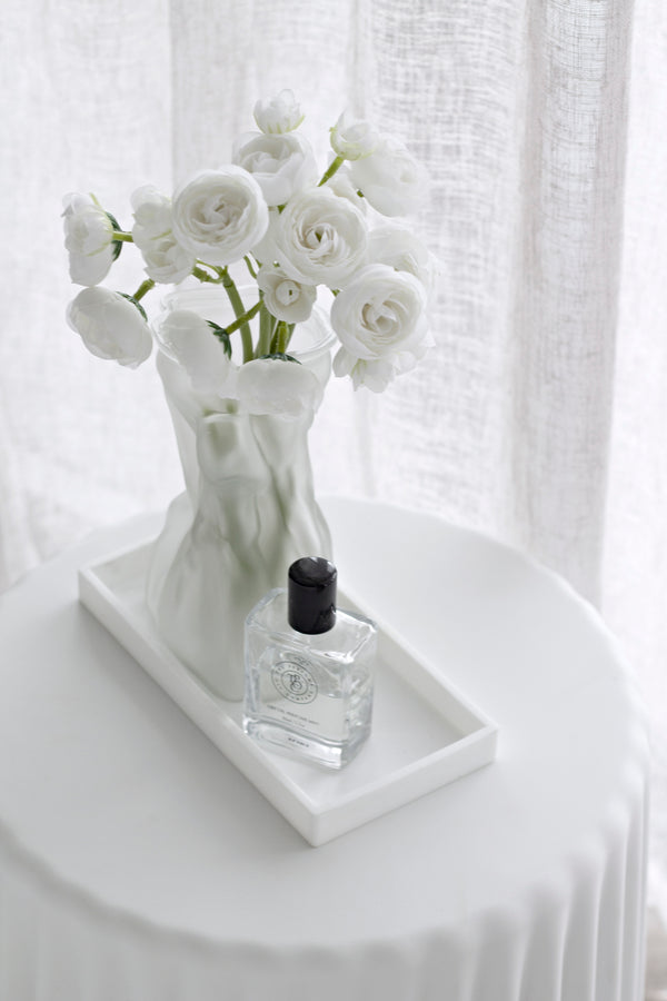 Clear glass perfume oil bottle styled on a white tray with fresh white ranunculus flowers in a vase, minimalist home setting.