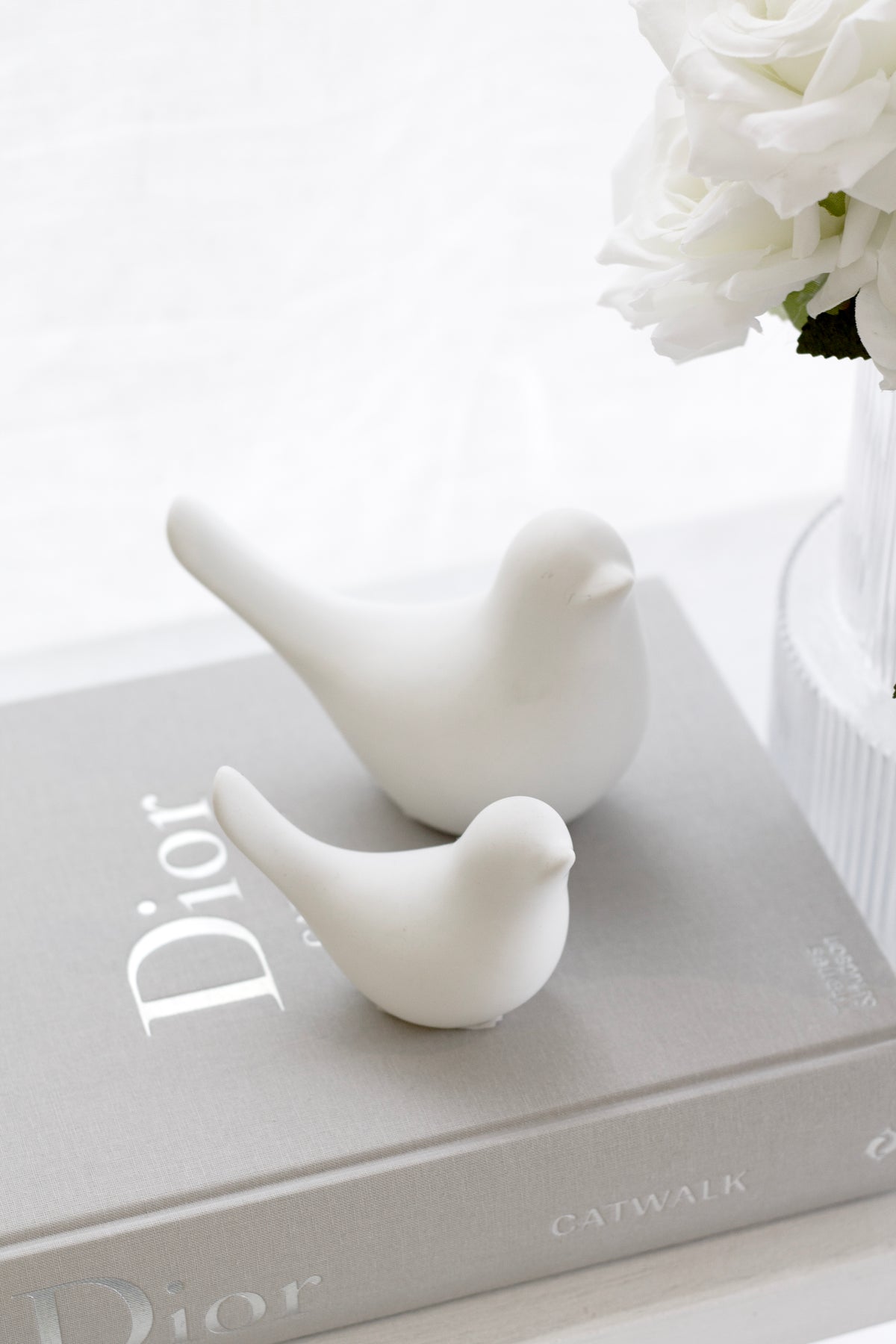 Two matte white ceramic doves on a grey Dior book beside white roses