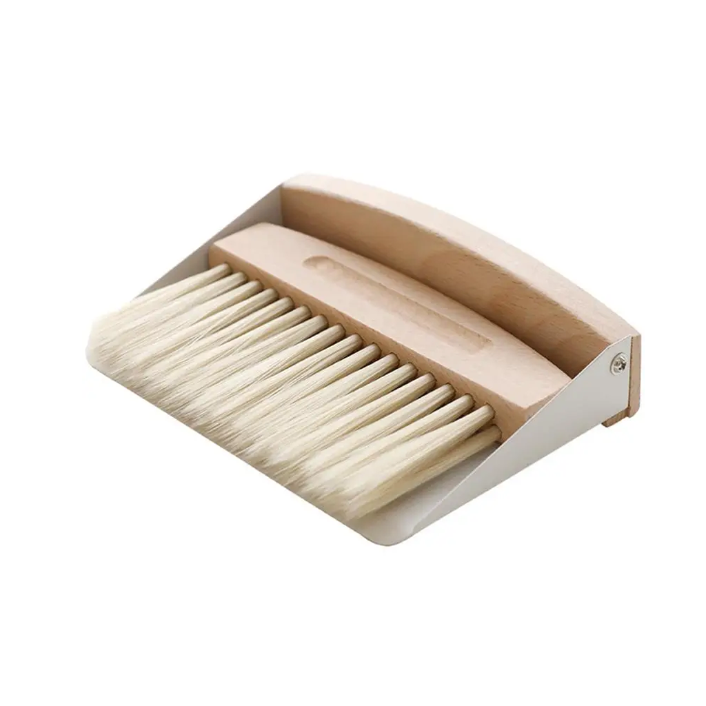 Wooden handled brush resting in its cream tabletop dustpan, shown as a set on white
