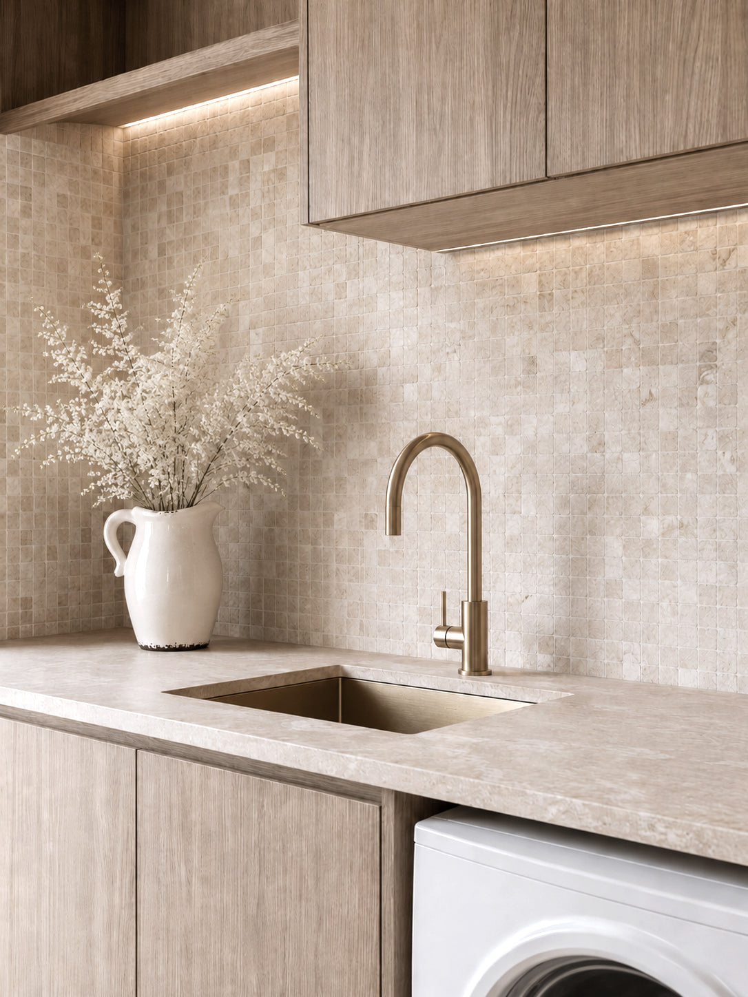Cream jug of fine dried stems on a stone benchtop in a pale timber scullery with brass tapware
