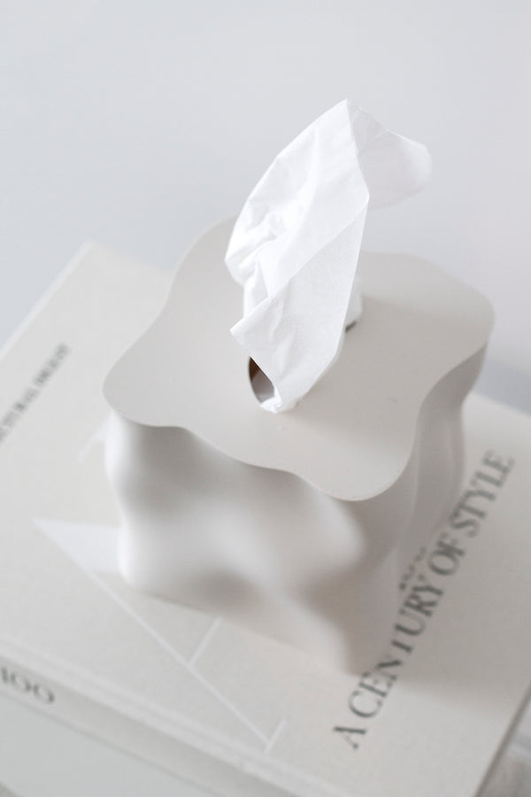 Sculptural wavy 3D-printed tissue box cover in matte white sitting on neutral coffee table books with a tissue pulled through.