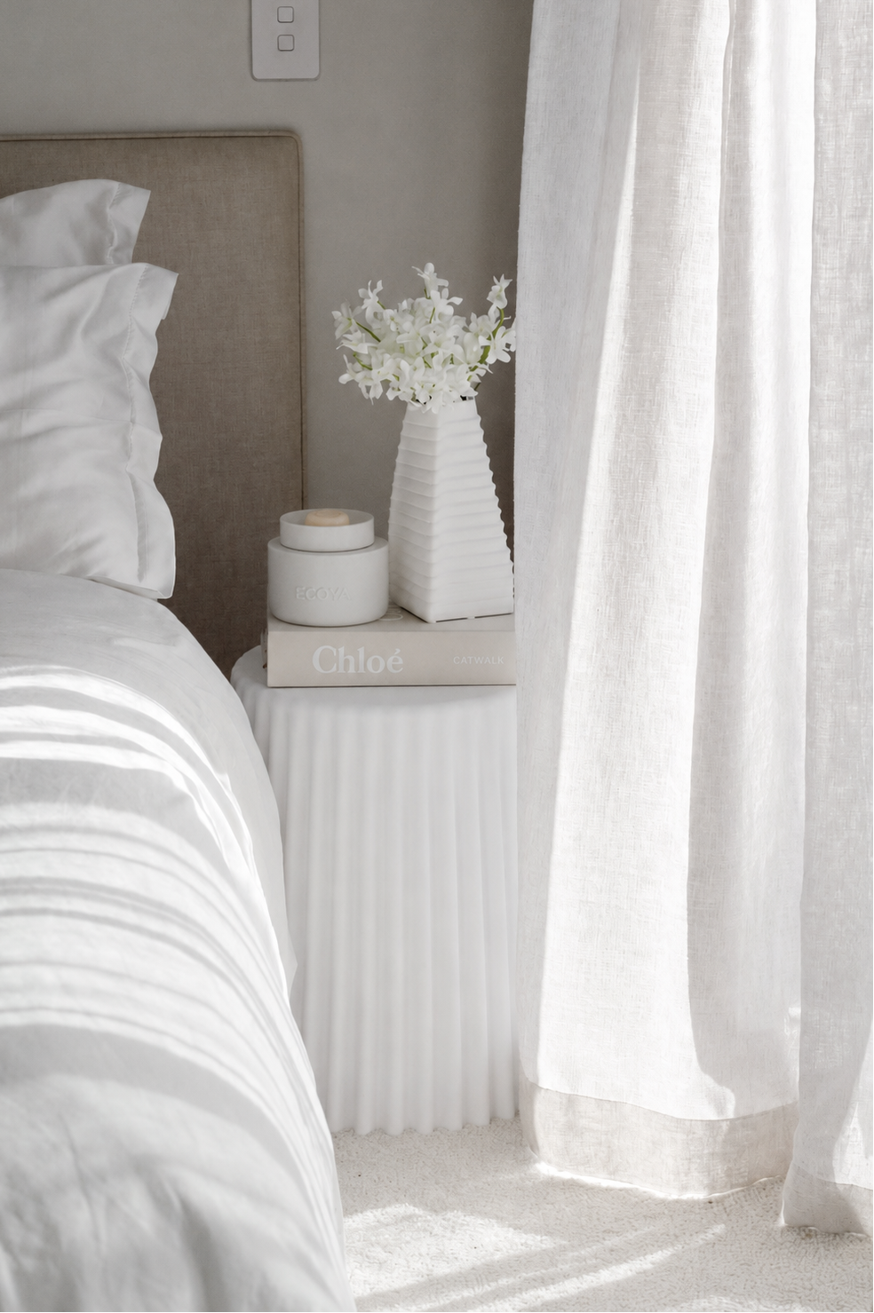 Small white ceramic vessel on a fluted bedside table beside a ribbed vase of white blossom