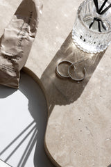 Ecoya Celebration diffuser in a ribbed clear glass bottle with black reeds, styled on a stone surface with jewellery hoops and a hand cream tube in soft sunlight