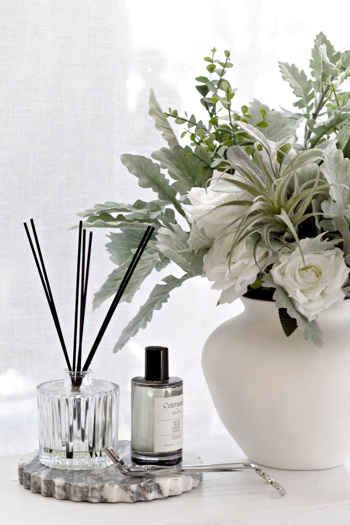 An elegant home decor vignette featuring the Ecoya Celebration Room Spray and matching diffuser styled on a chic, grey marble tray. The home fragrance set is placed next to a large white vase with a lush arrangement of white roses and silvery-green foliage, creating a sophisticated, serene, and luxurious atmosphere.