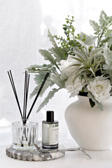An elegant home decor vignette featuring the Ecoya Celebration Room Spray and matching diffuser styled on a chic, grey marble tray. The home fragrance set is placed next to a large white vase with a lush arrangement of white roses and silvery-green foliage, creating a sophisticated, serene, and luxurious atmosphere.