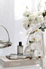 Black Ecoya Celebration Room Spray styled with white orchids, books, and a silver decorative object on a bright console table.