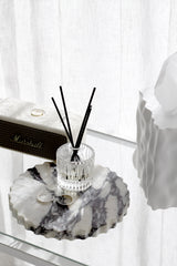 Glass reed diffuser with black reeds on a scalloped marble tray beside jewellery, in soft window light.