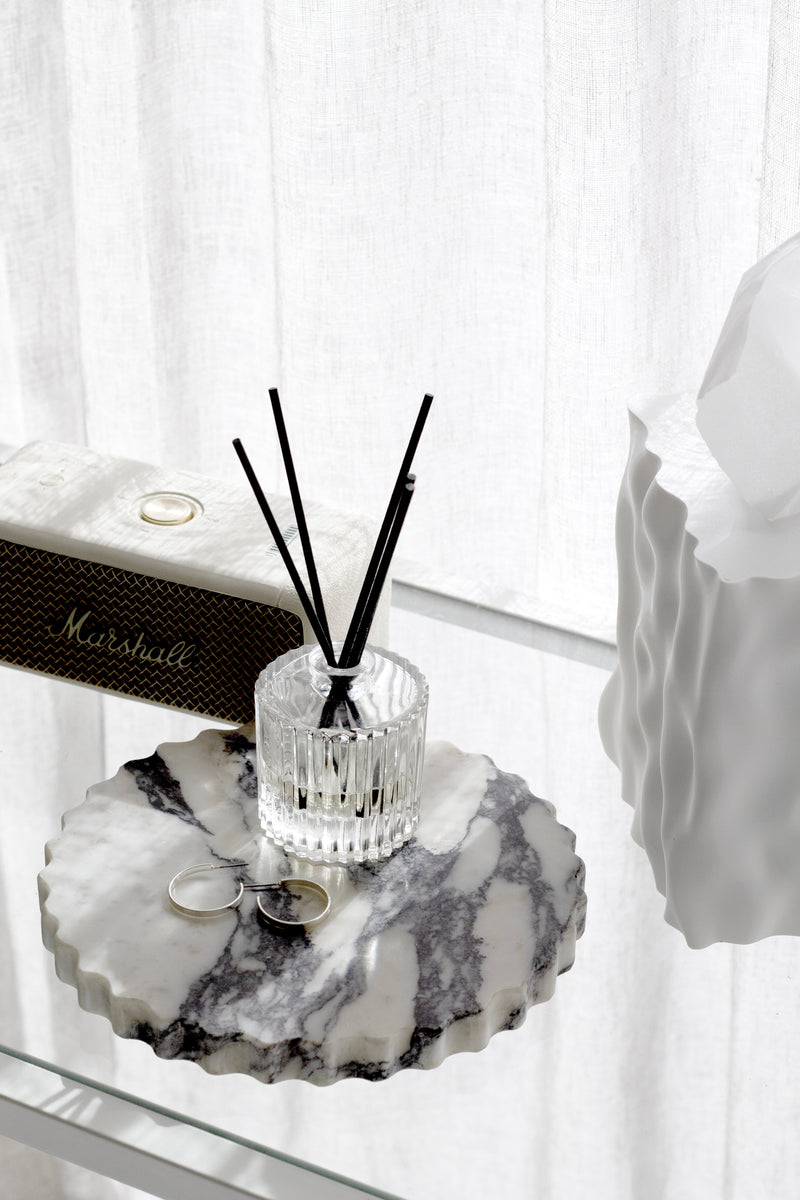 Glass reed diffuser with black reeds on a scalloped marble tray beside jewellery, in soft window light.