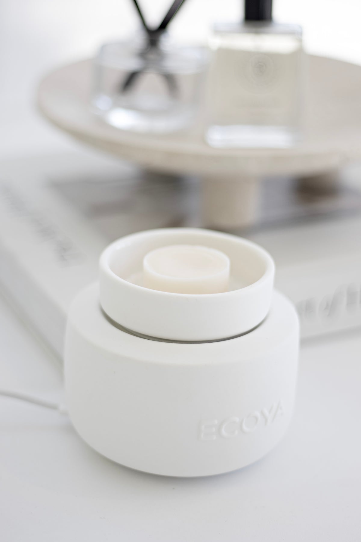 ECOYA Electric Wax Melter in white ceramic on a book, plugged in, with a blurred reed diffuser styled behind.