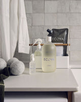 Ecoya fragranced linen spray 100ml styled in a laundry setting with Ecoya bottle and refill pouch, spray bottle, dryer balls and folded linens against grey tiled wall