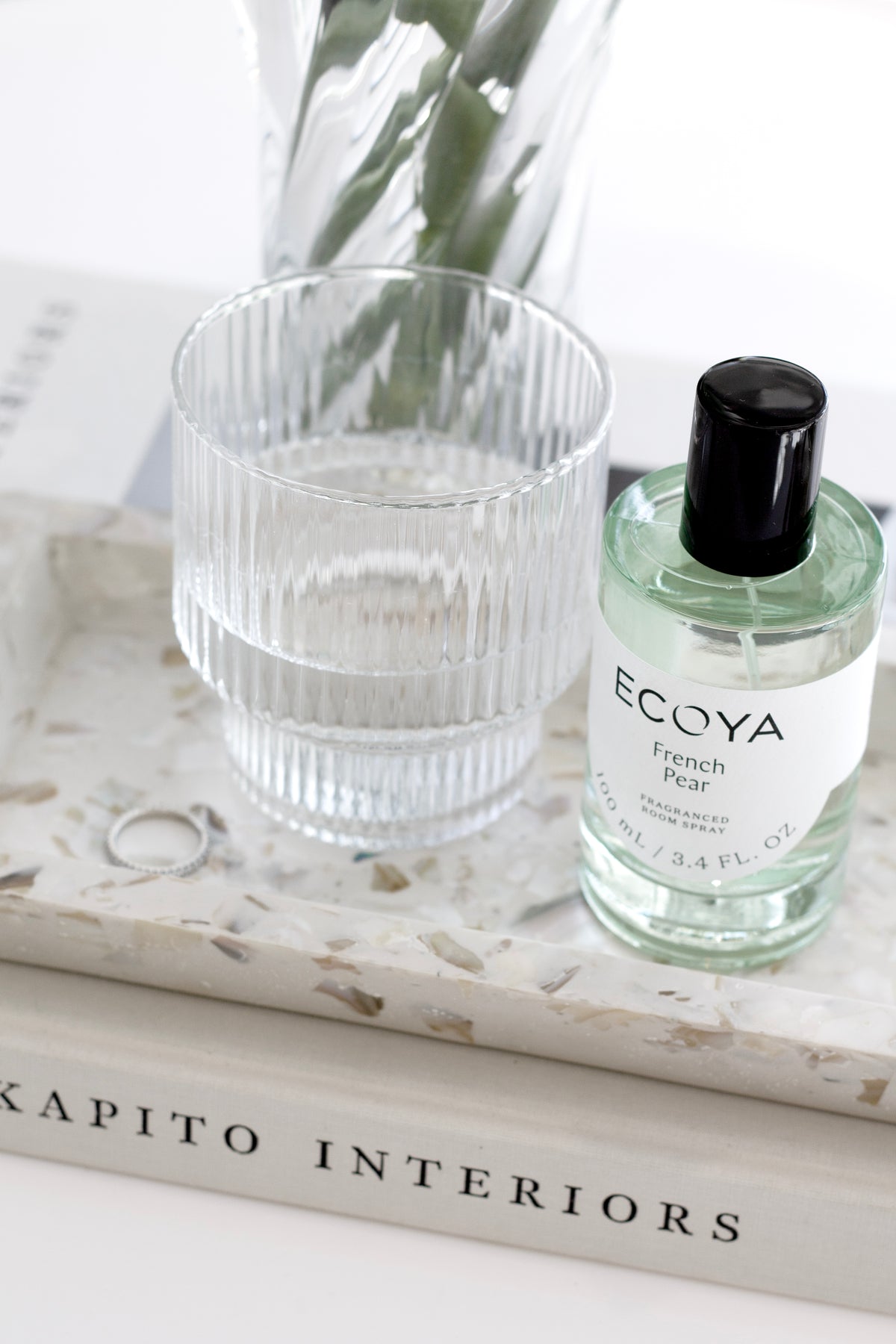 Green-tinted Ecoya room spray on a terrazzo tray beside a ribbed glass vase on a book