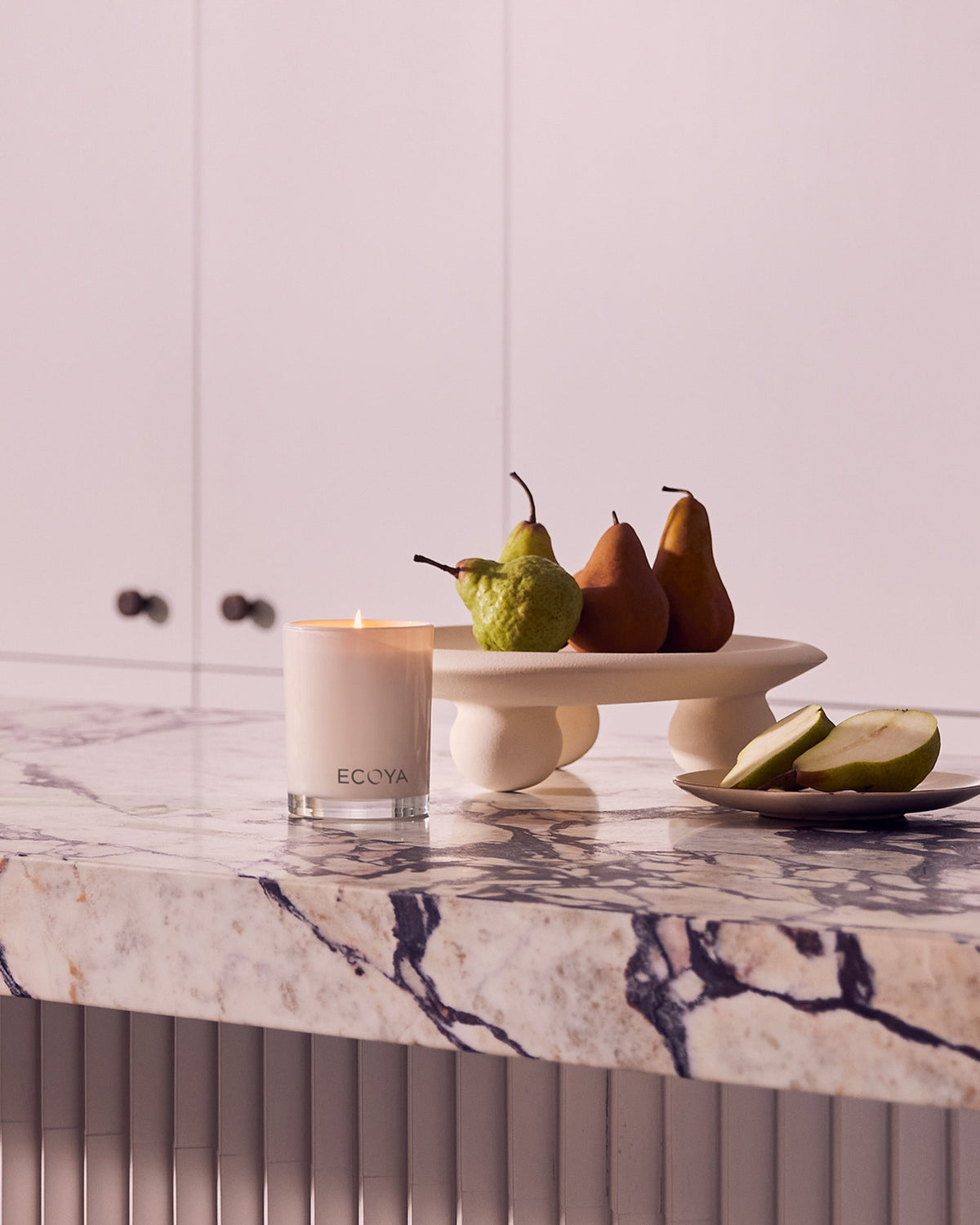 A lit Ecoya Madison candle on a marble benchtop beside pears on a pedestal stand and sliced pear on a plate
