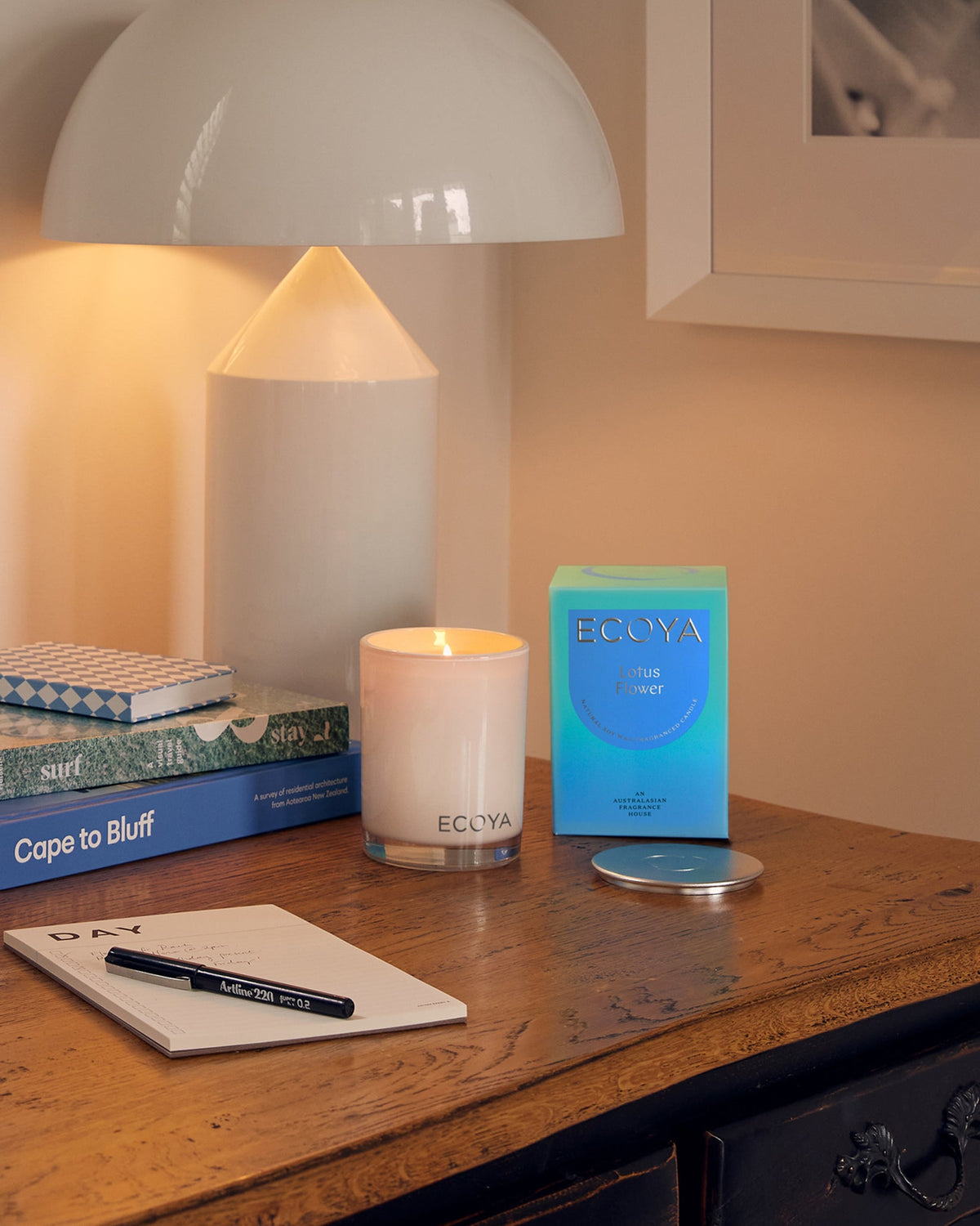 A lit Ecoya Madison candle on a timber desk beside its blue gift box, a dome lamp and a notebook