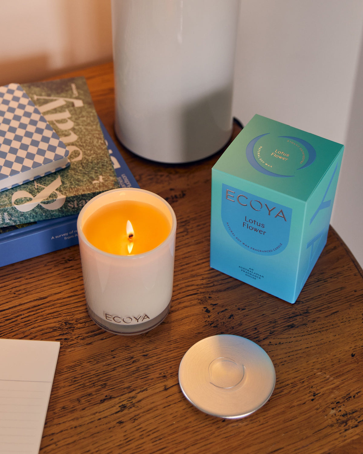 A lit Ecoya Madison candle on a timber bedside table beside its blue gift box and silver lid, with books alongside