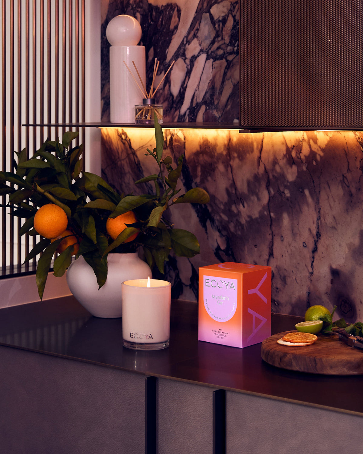 A lit Ecoya Madison candle beside its coral gift box on a dark console, with a vase of mandarins on the branch alongside