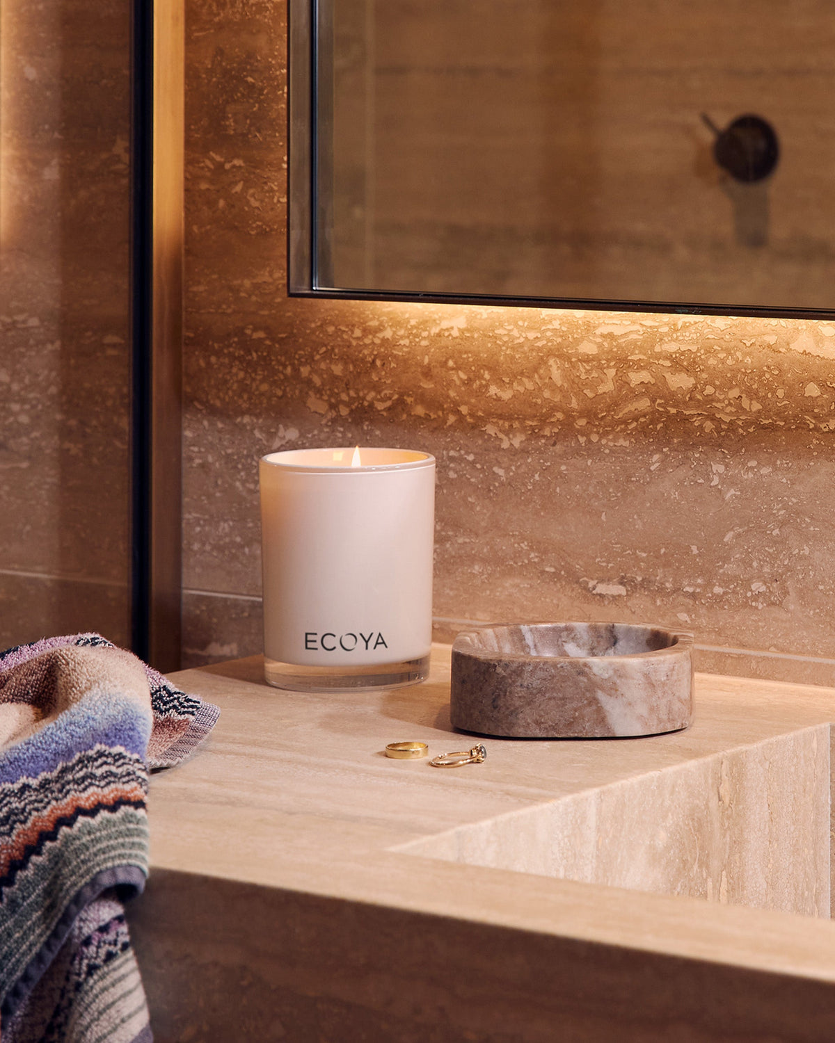 A lit Ecoya Madison candle on a travertine vanity beside a stone dish, with warm backlit stone walls behind