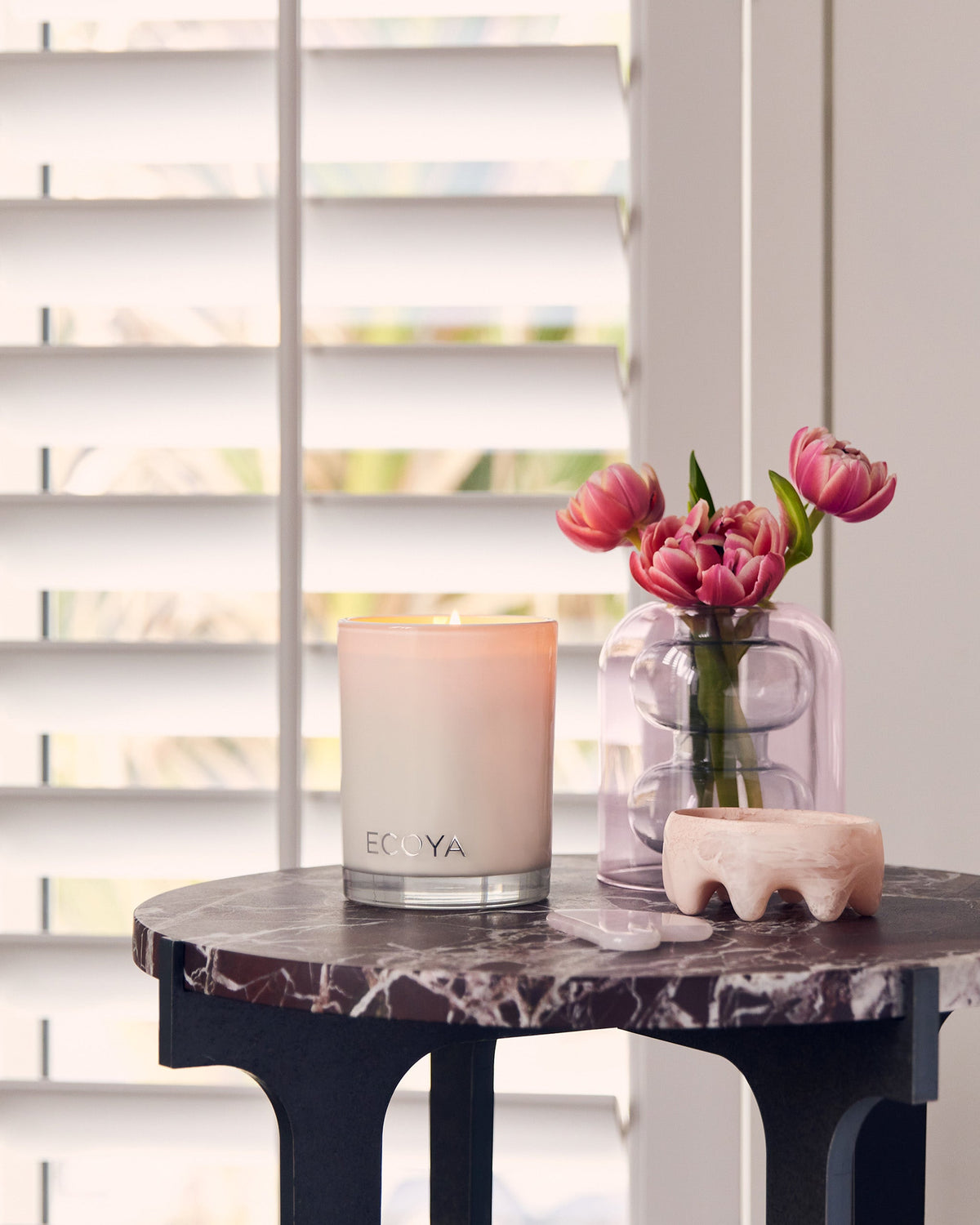 Ecoya Madison candle on a dark side table beside pink roses by white shutters